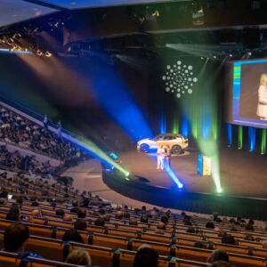 Stort auditorium med scen, stor skärm och åhörare; presentatörer står på scenen.