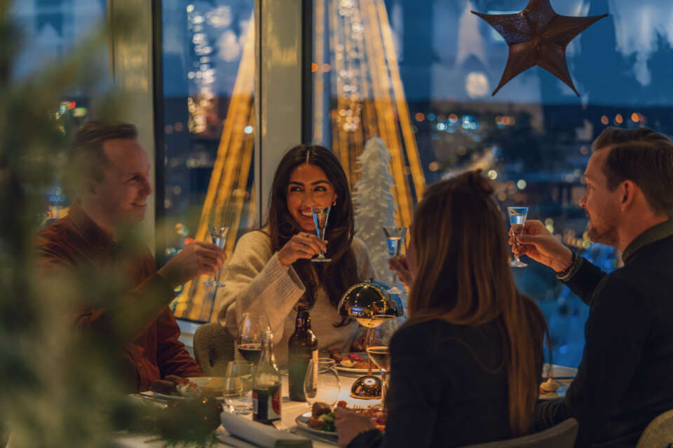 Julbord med fyra personer som skålar runt ett bord, med juldekorationer i bakgrunden.