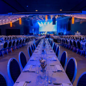 Storsal dukad för middag med långbord och stolar, blåljusbelyst scen i bakgrunden.